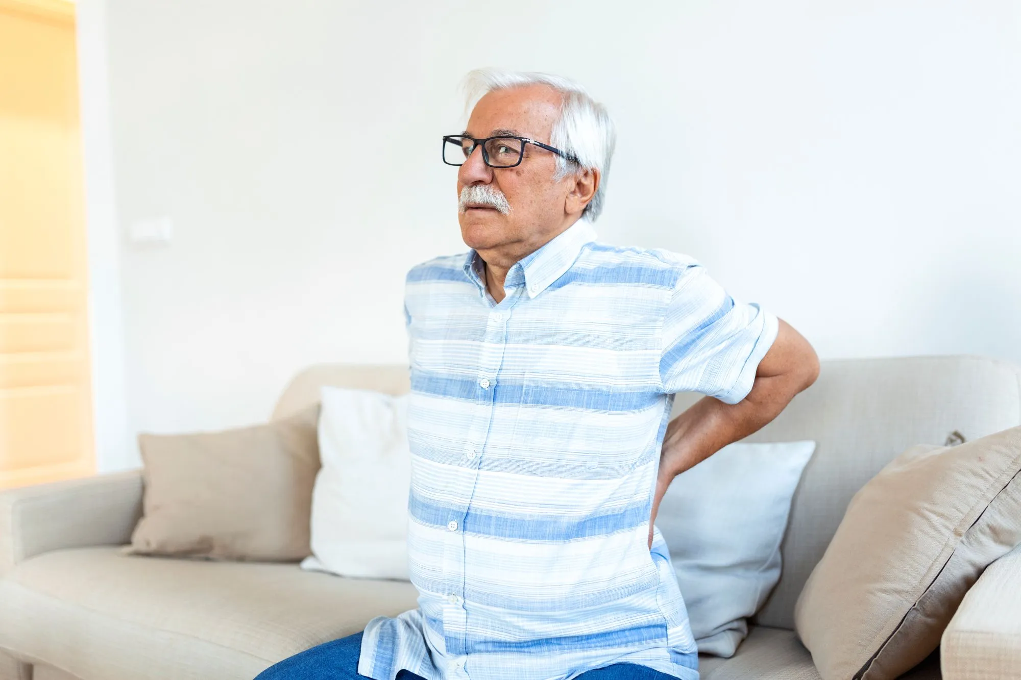Elderly man sitting on a couch, clutching his lower back with a pained expression